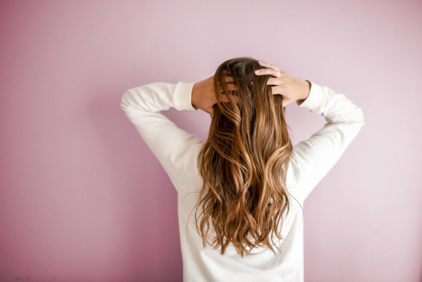 A woman runs her hands through glossy, healthy hair against a soft rose backdrop